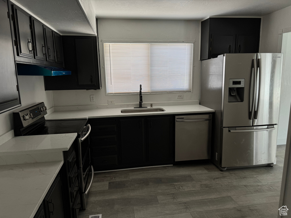 Kitchen featuring stainless steel appliances, dark cabinets, under cabinet range hood, light wood-style flooring, and light stone countertops