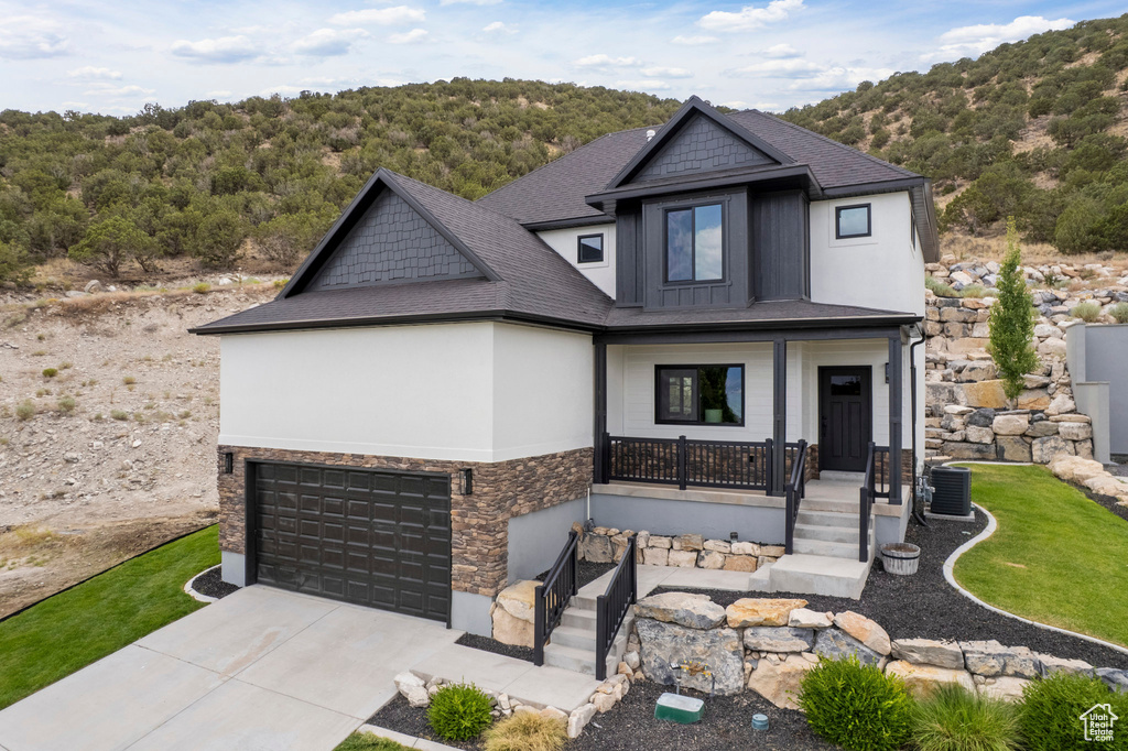 Craftsman-style house with stone siding, covered porch, concrete driveway, an attached garage, and a view of trees
