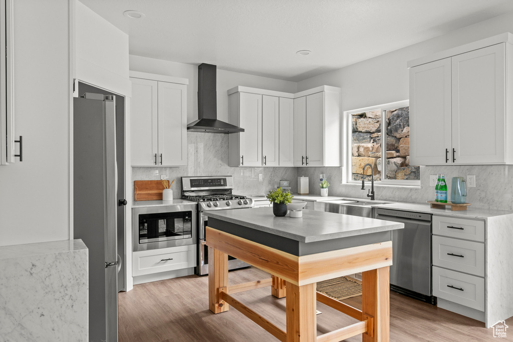 Kitchen with appliances with stainless steel finishes, wall chimney range hood, white cabinetry, decorative backsplash, and light wood-type flooring