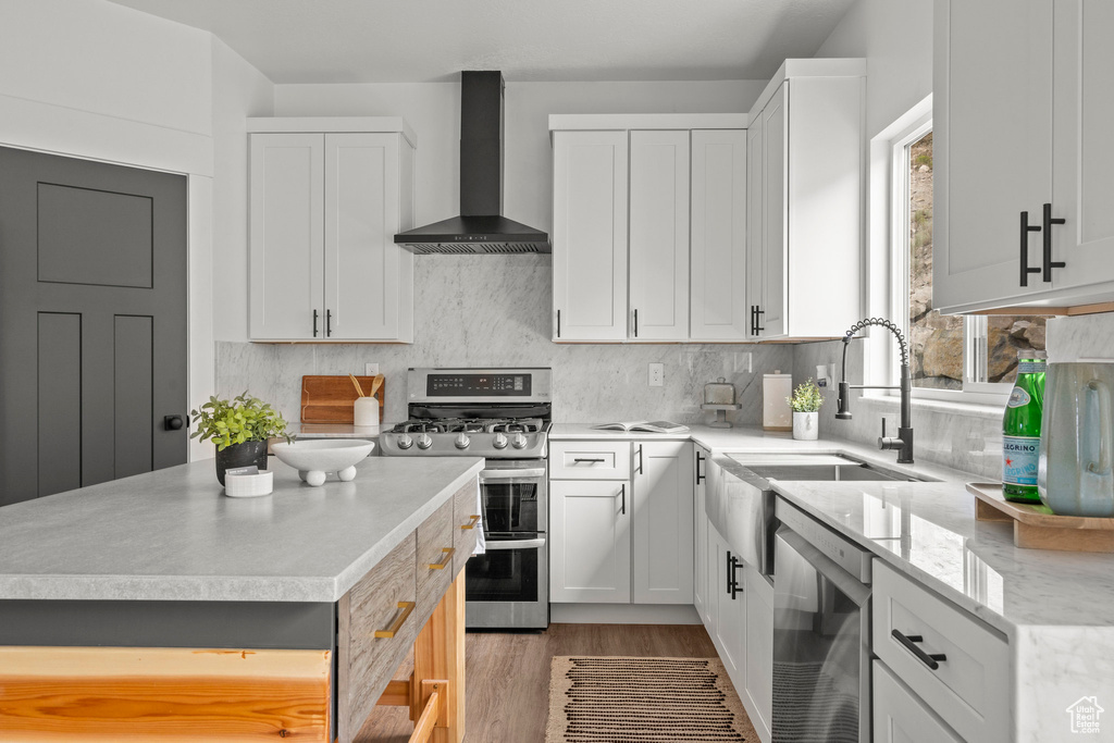Kitchen featuring wall chimney exhaust hood, appliances with stainless steel finishes, light wood-type flooring, backsplash, and white cabinetry