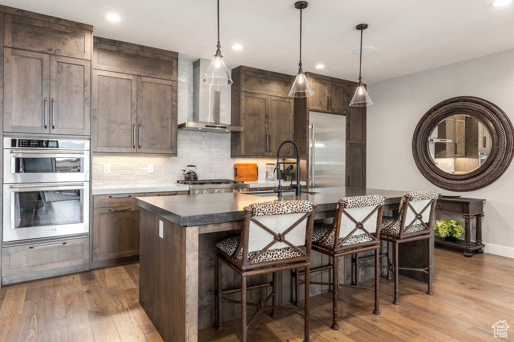 Kitchen with dark wood-style flooring, a kitchen island with sink, decorative backsplash, wall chimney range hood, and recessed lighting