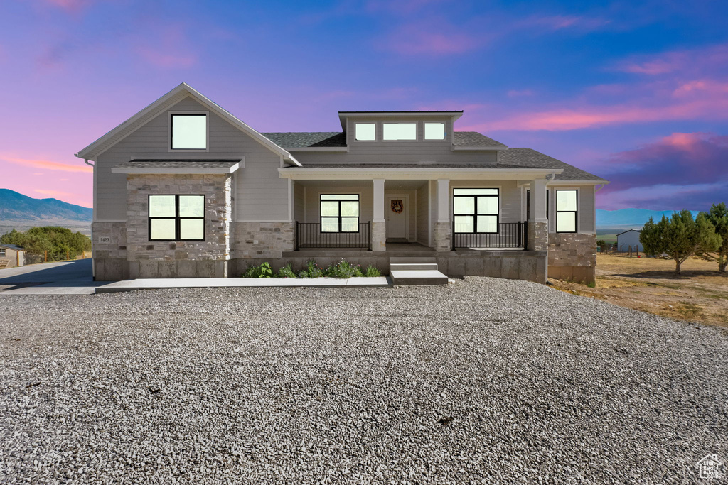 View of front of home with stone siding, covered porch, and roof with shingles