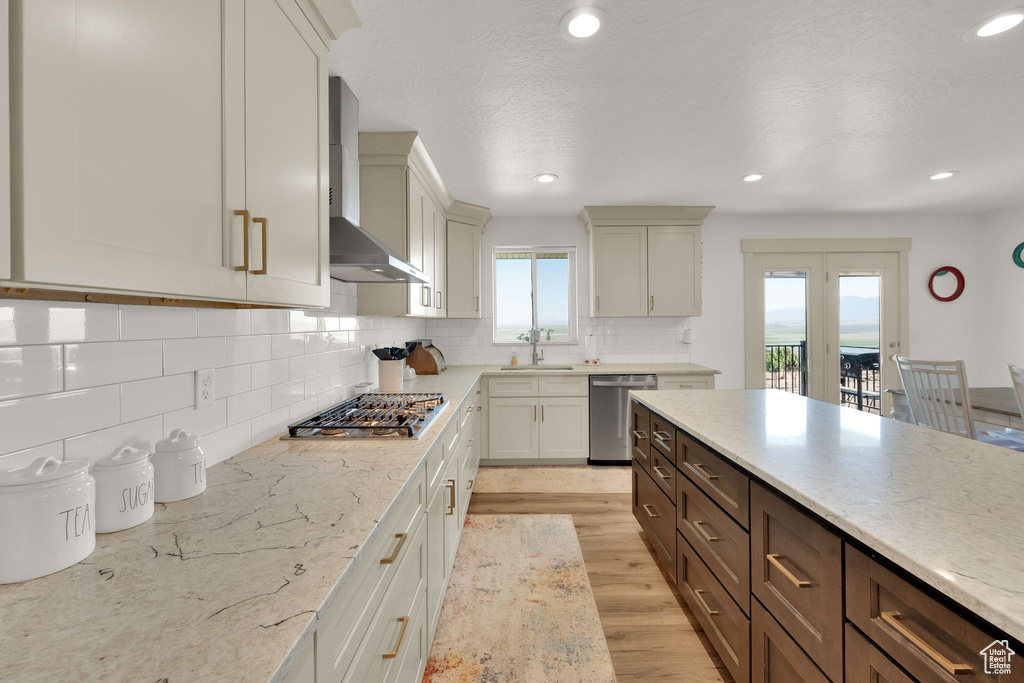 Kitchen featuring stainless steel appliances, wall chimney exhaust hood, decorative backsplash, light wood-style floors, and light stone counters