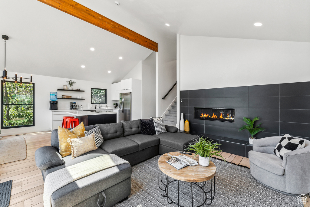 Living room with light wood finished floors, a tiled fireplace, beam ceiling, recessed lighting, and stairs