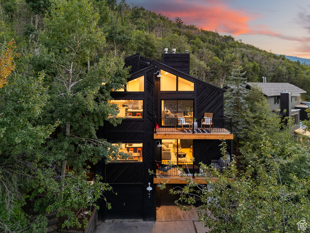 View of front of home with a balcony, a chimney, a forest view, concrete driveway, and a garage