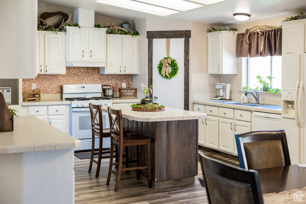 Kitchen with white appliances, under cabinet range hood, white cabinets, wood finished floors, and backsplash