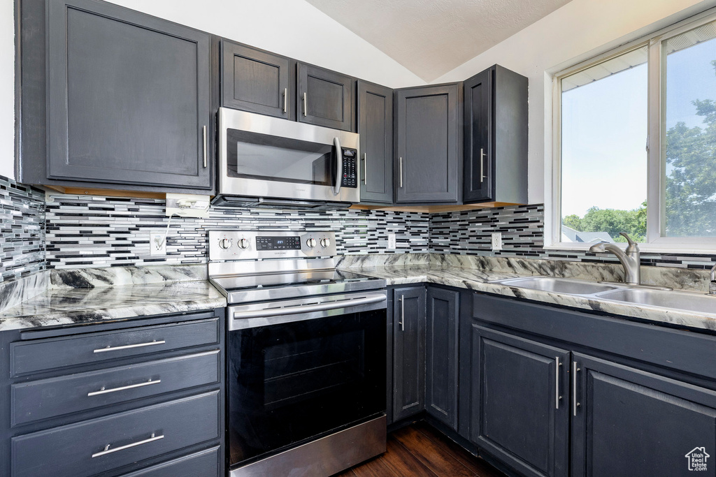 Kitchen with appliances with stainless steel finishes, lofted ceiling, tasteful backsplash, light stone countertops, and dark wood finished floors