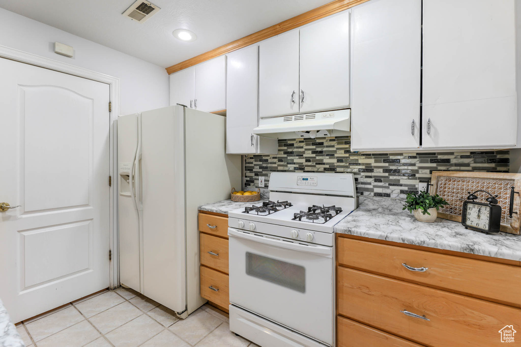 Kitchen featuring white appliances, under cabinet range hood, backsplash, light tile patterned flooring, and white cabinetry