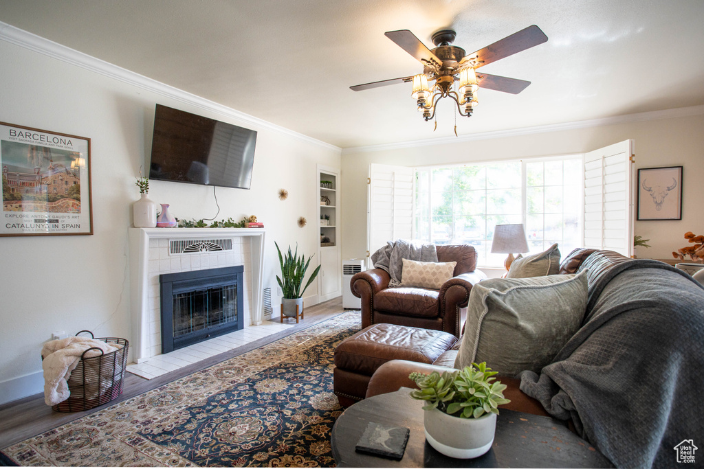 Living room featuring crown molding, a tiled fireplace, ceiling fan, wood finished floors, and radiator heating unit