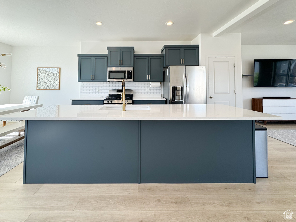 Kitchen with stainless steel appliances, light countertops, a large island with sink, tasteful backsplash, and light wood-style floors