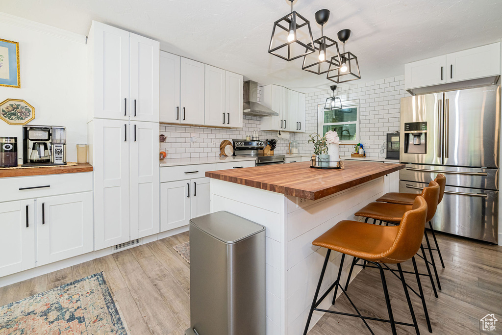 Kitchen featuring butcher block counters, stainless steel appliances, wall chimney exhaust hood, white cabinetry, and a kitchen bar