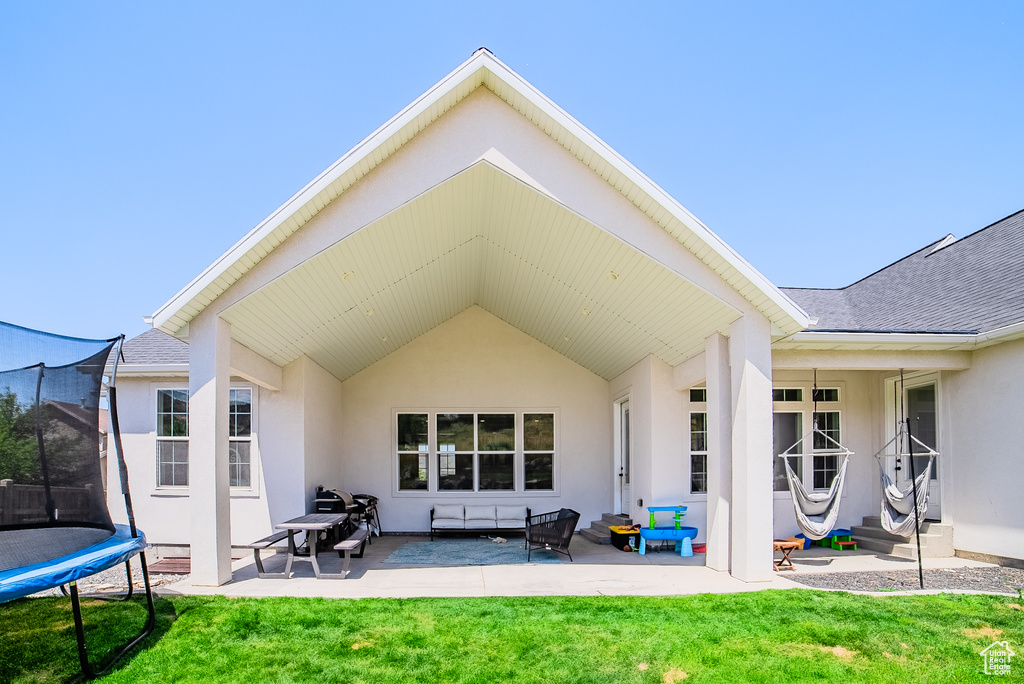 Rear view of house with a trampoline, a patio, stucco siding, a yard, and roof with shingles