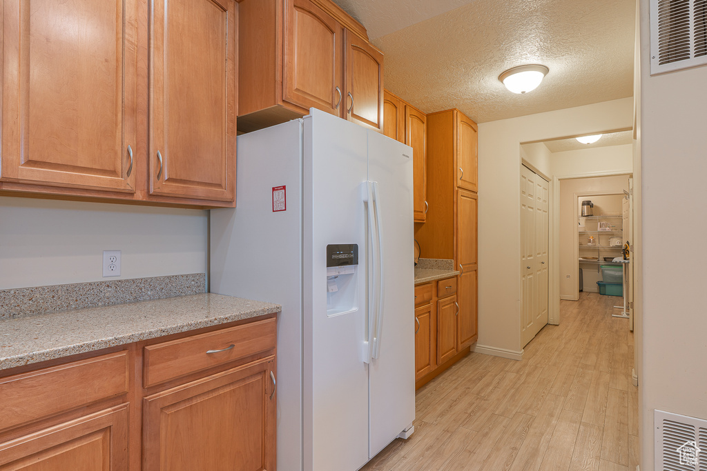 Kitchen featuring white refrigerator with ice dispenser, light wood finished floors, a textured ceiling, brown cabinets, and light stone countertops