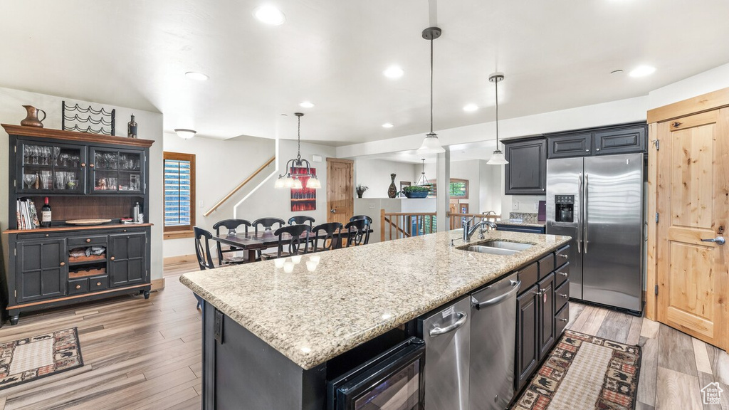 Kitchen with stainless steel appliances, a kitchen island with sink, light wood-style flooring, light stone counters, and hanging light fixtures