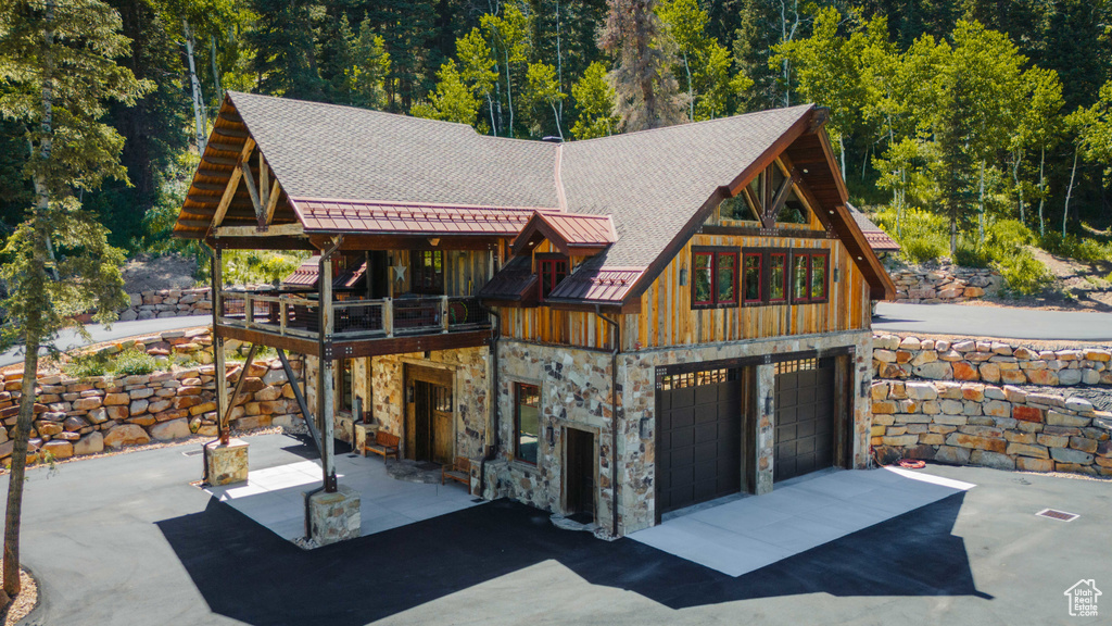 View of front facade featuring stone siding, a standing seam roof, driveway, a balcony, and a view of trees