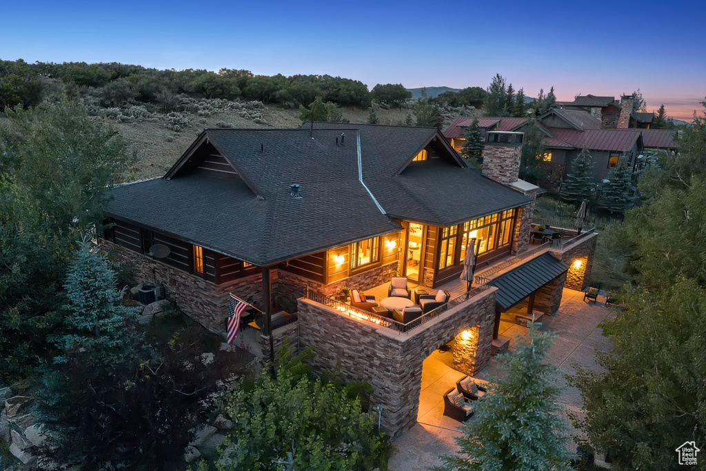 Back of property at dusk with a shingled roof, stone siding, a chimney, and a patio area