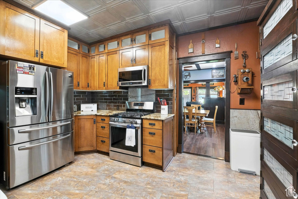 Kitchen featuring appliances with stainless steel finishes, brown cabinetry, an ornate ceiling, light stone counters, and glass insert cabinets