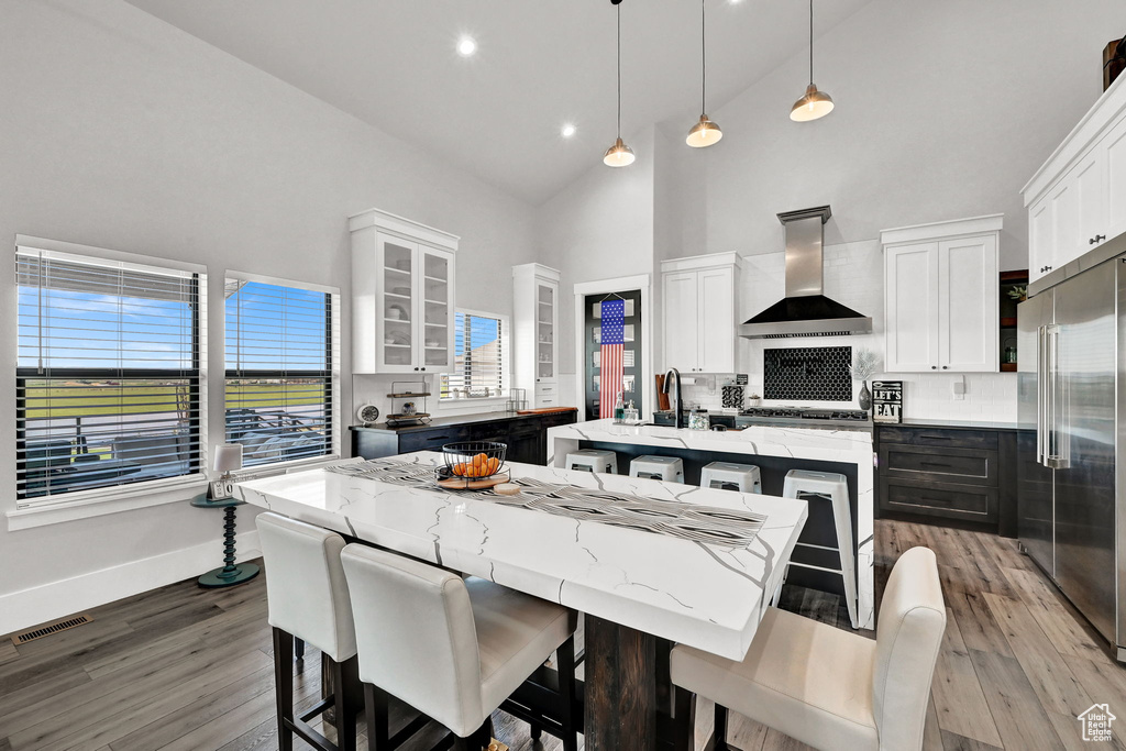 Kitchen featuring wall chimney range hood, high vaulted ceiling, an island with sink, stainless steel appliances, and a kitchen bar