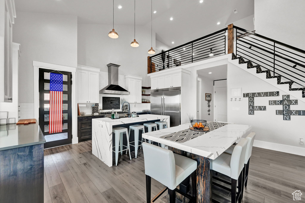Kitchen with wall chimney range hood, high vaulted ceiling, built in refrigerator, a center island with sink, and tasteful backsplash