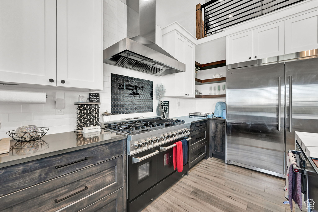 Kitchen with wall chimney range hood, high quality appliances, white cabinets, tasteful backsplash, and light wood-style flooring
