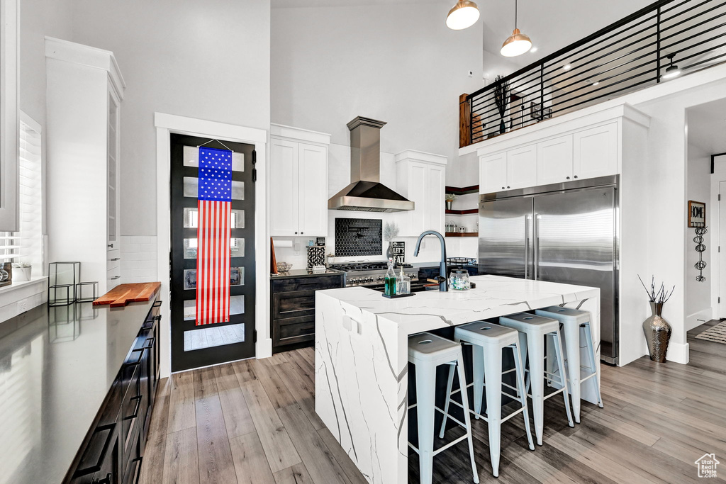 Kitchen featuring wall chimney exhaust hood, a high ceiling, built in fridge, light wood-style flooring, and white cabinets