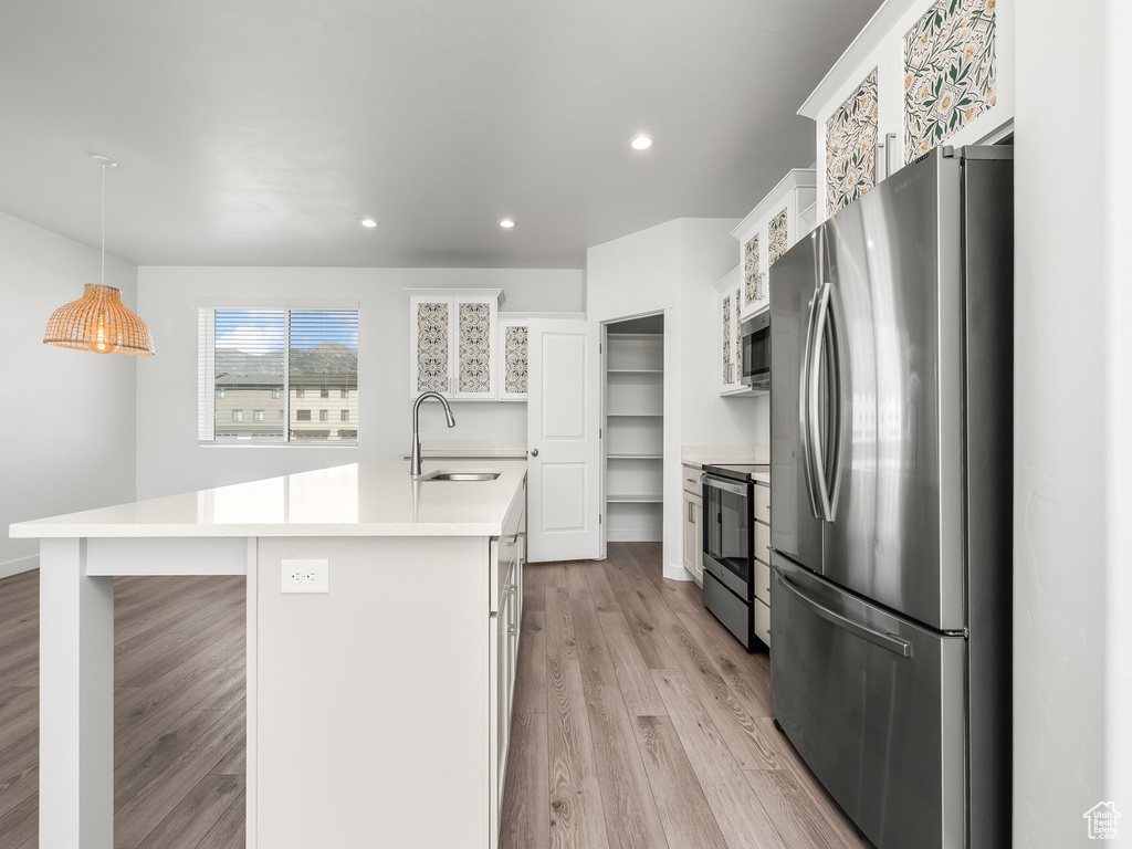 Kitchen with appliances with stainless steel finishes, light wood finished floors, a kitchen island with sink, light countertops, and white cabinetry