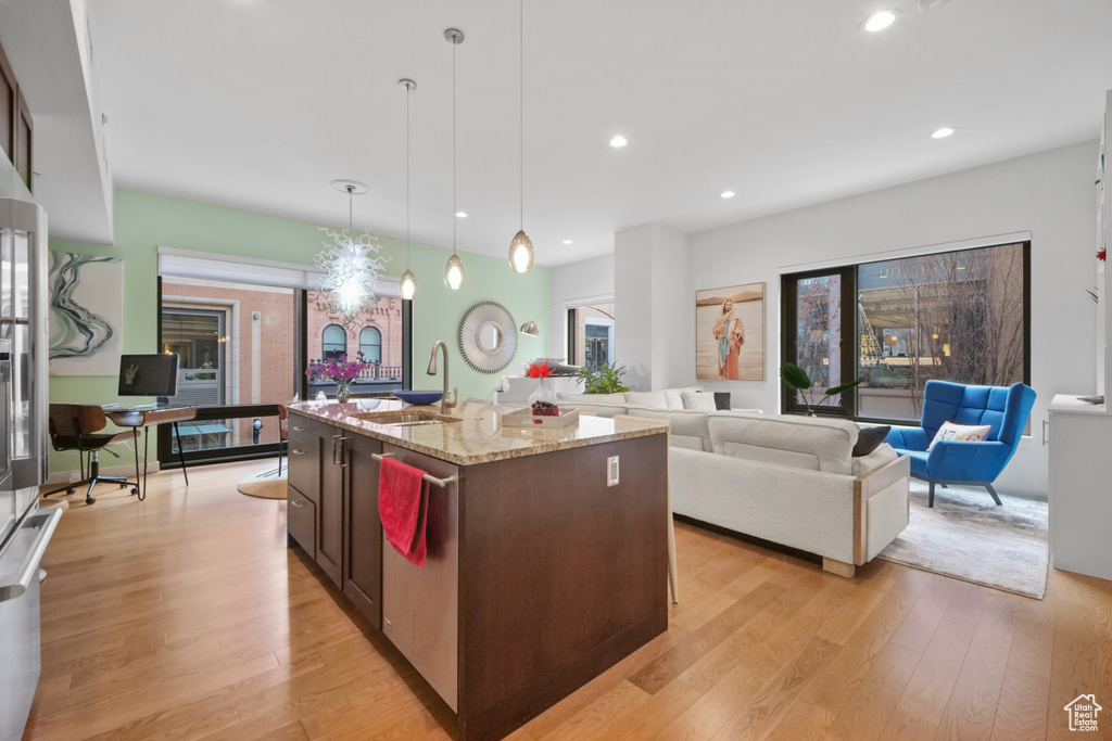 Kitchen with dishwasher, light wood-type flooring, light stone counters, pendant lighting, and a center island with sink