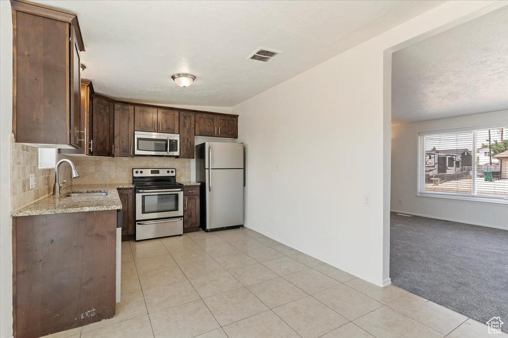 Kitchen with appliances with stainless steel finishes, decorative backsplash, dark brown cabinets, light stone countertops, and light tile patterned flooring