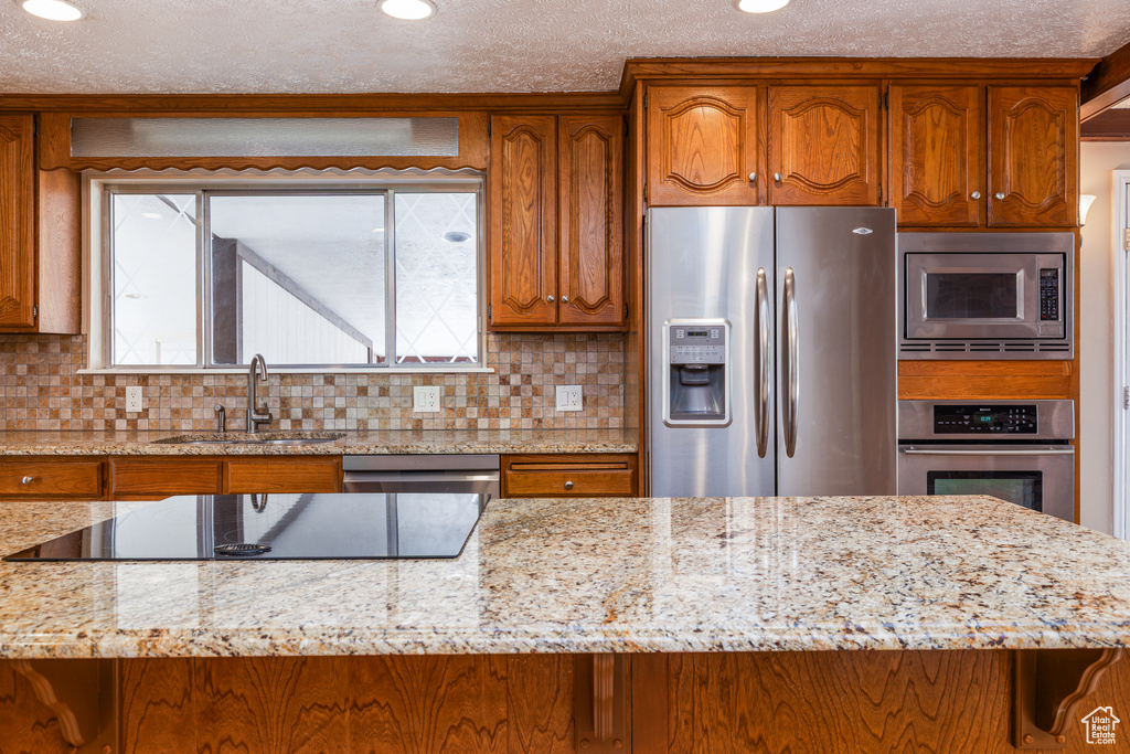 Kitchen with appliances with stainless steel finishes, brown cabinetry, backsplash, light stone counters, and recessed lighting