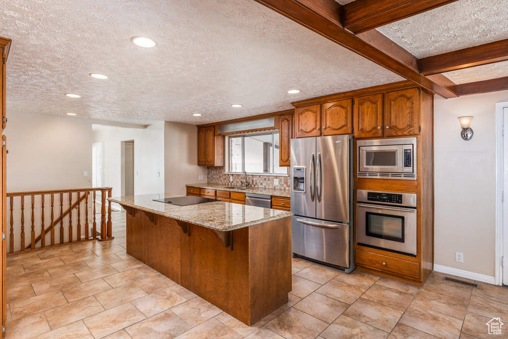 Kitchen with appliances with stainless steel finishes, brown cabinets, light stone countertops, a textured ceiling, and tasteful backsplash