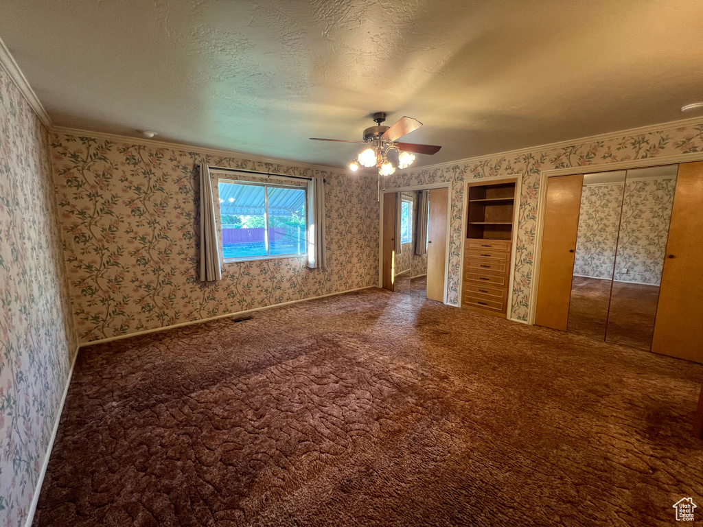 Unfurnished bedroom featuring wallpapered walls, crown molding, multiple closets, carpet, and a ceiling fan