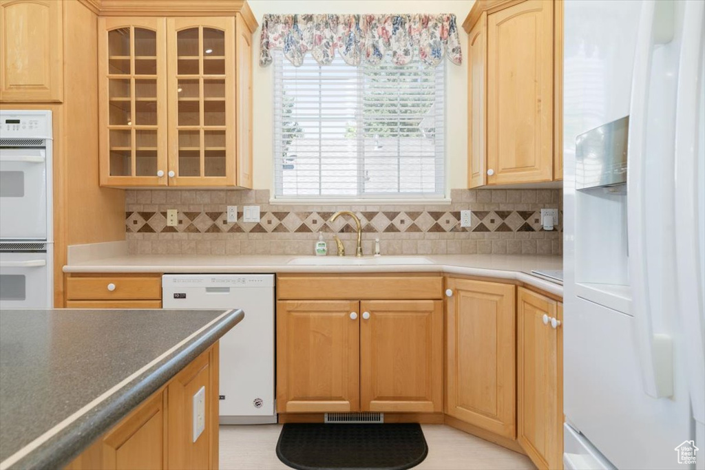 Kitchen with white appliances, backsplash, light brown cabinetry, and glass insert cabinets