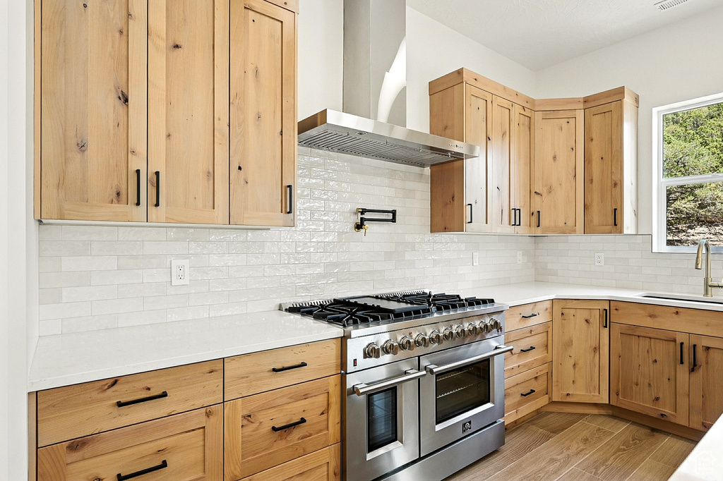 Kitchen with light brown cabinetry, range with two ovens, wall chimney range hood, and light countertops