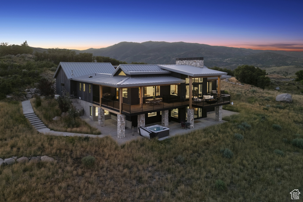 Back of house at dusk with stone siding, a chimney, a patio, a deck with mountain view, and a metal roof