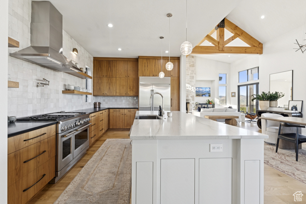 Kitchen with premium appliances, wall chimney exhaust hood, light wood finished floors, decorative backsplash, and a center island with sink