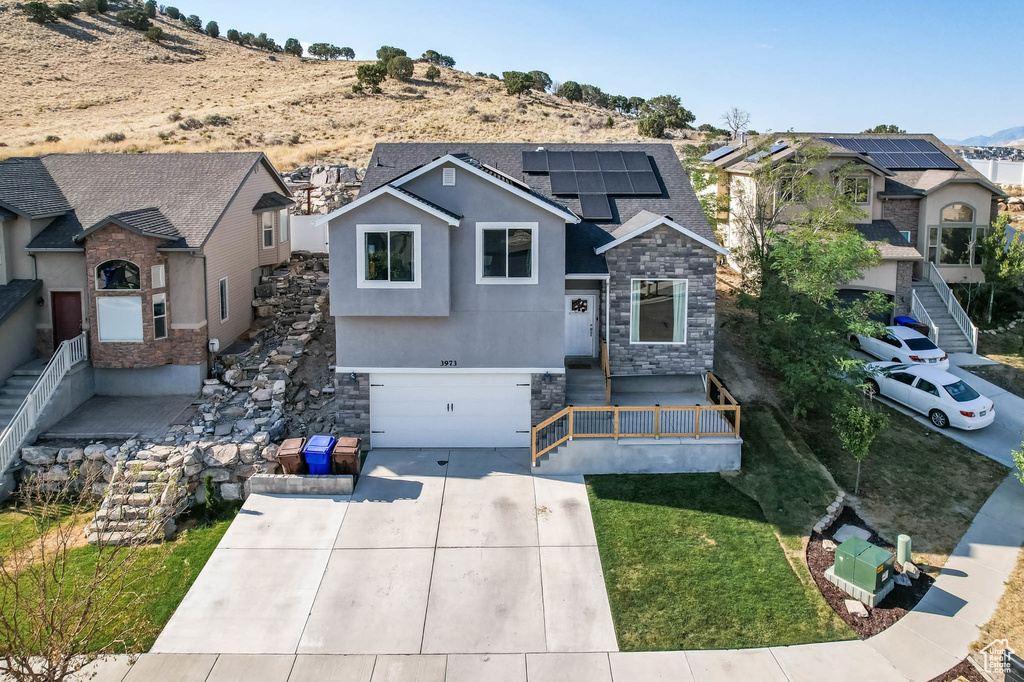 View of front of property with stone siding, a garage, driveway, roof mounted solar panels, and roof with shingles