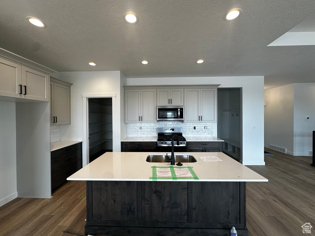 Kitchen featuring gray cabinets, appliances with stainless steel finishes, dark wood-style floors, decorative backsplash, and a kitchen island with sink
