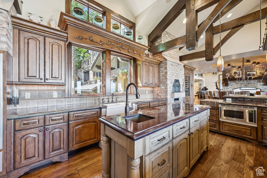Kitchen with stainless steel microwave, a kitchen island with sink, backsplash, dark wood finished floors, and beamed ceiling