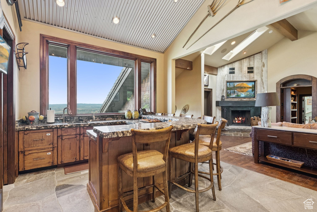 Kitchen featuring beam ceiling, dark stone counters, a kitchen bar, a stone fireplace, and brown cabinetry
