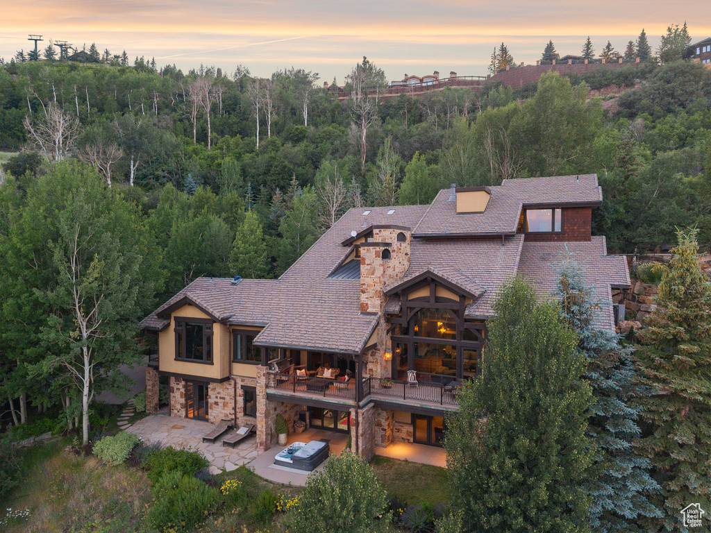 Rear view of house with stone siding, a balcony, a patio, a view of trees, and a shingled roof