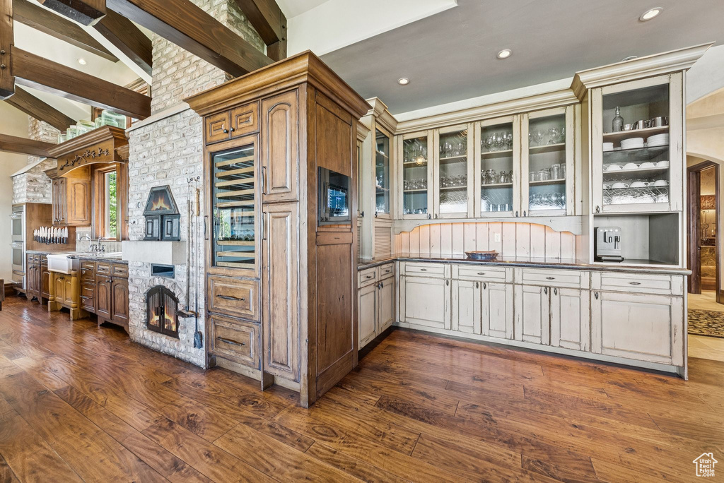 Kitchen featuring dark wood-type flooring, glass insert cabinets, beam ceiling, dark countertops, and a fireplace