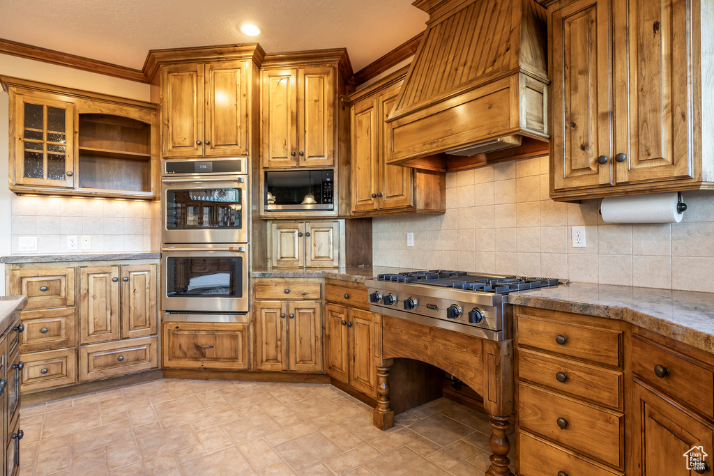 Kitchen featuring stainless steel appliances, premium range hood, tasteful backsplash, brown cabinets, and open shelves