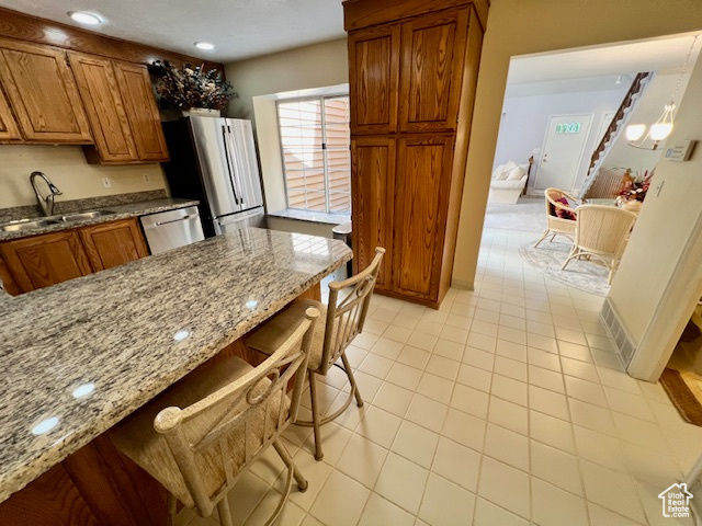Kitchen featuring appliances with stainless steel finishes, brown cabinets, light stone countertops, light tile patterned floors, and a breakfast bar