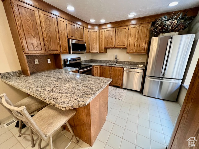 Kitchen featuring appliances with stainless steel finishes, a peninsula, brown cabinets, light stone counters, and light tile patterned floors