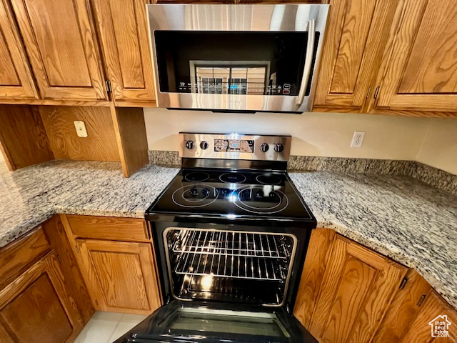 Kitchen featuring stainless steel microwave, black range with electric cooktop, brown cabinets, and light stone counters