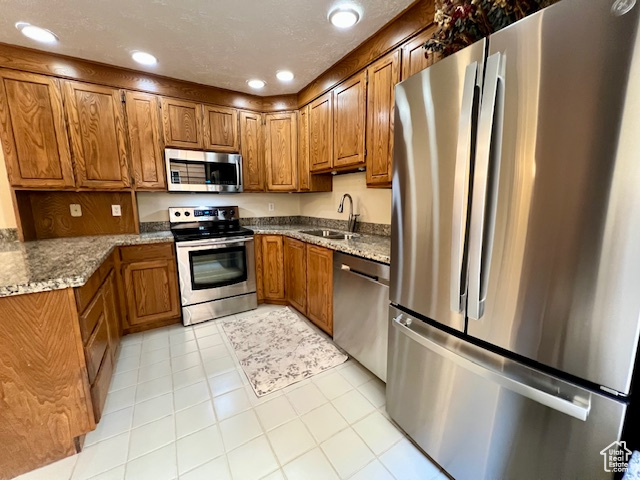 Kitchen featuring appliances with stainless steel finishes, brown cabinetry, dark stone counters, light tile patterned floors, and recessed lighting