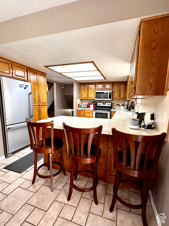 Kitchen featuring a textured ceiling, brown cabinets, light countertops, stainless steel appliances, and a peninsula