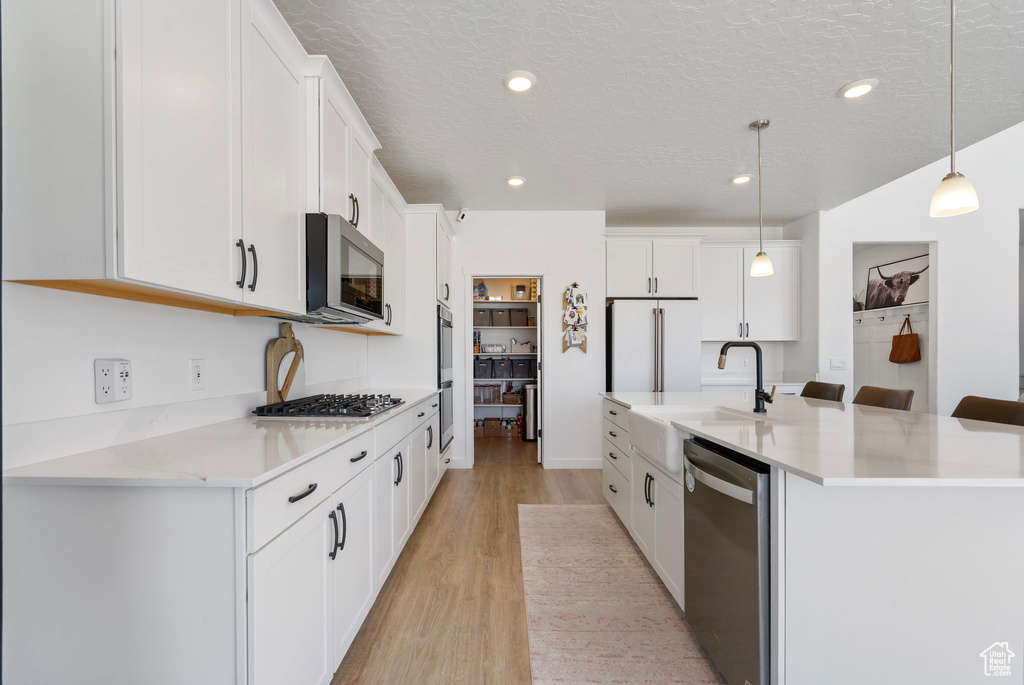 Kitchen with appliances with stainless steel finishes, light wood-style floors, a spacious island, white cabinets, and pendant lighting