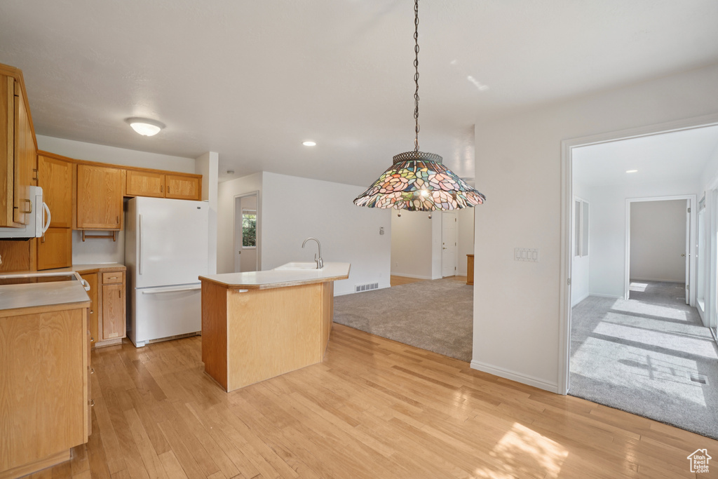 Kitchen with freestanding refrigerator, light wood-type flooring, a center island with sink, light countertops, and decorative light fixtures