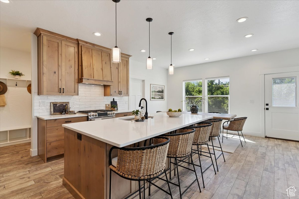 Kitchen with light wood-style flooring, light countertops, decorative backsplash, a kitchen island with sink, and custom range hood